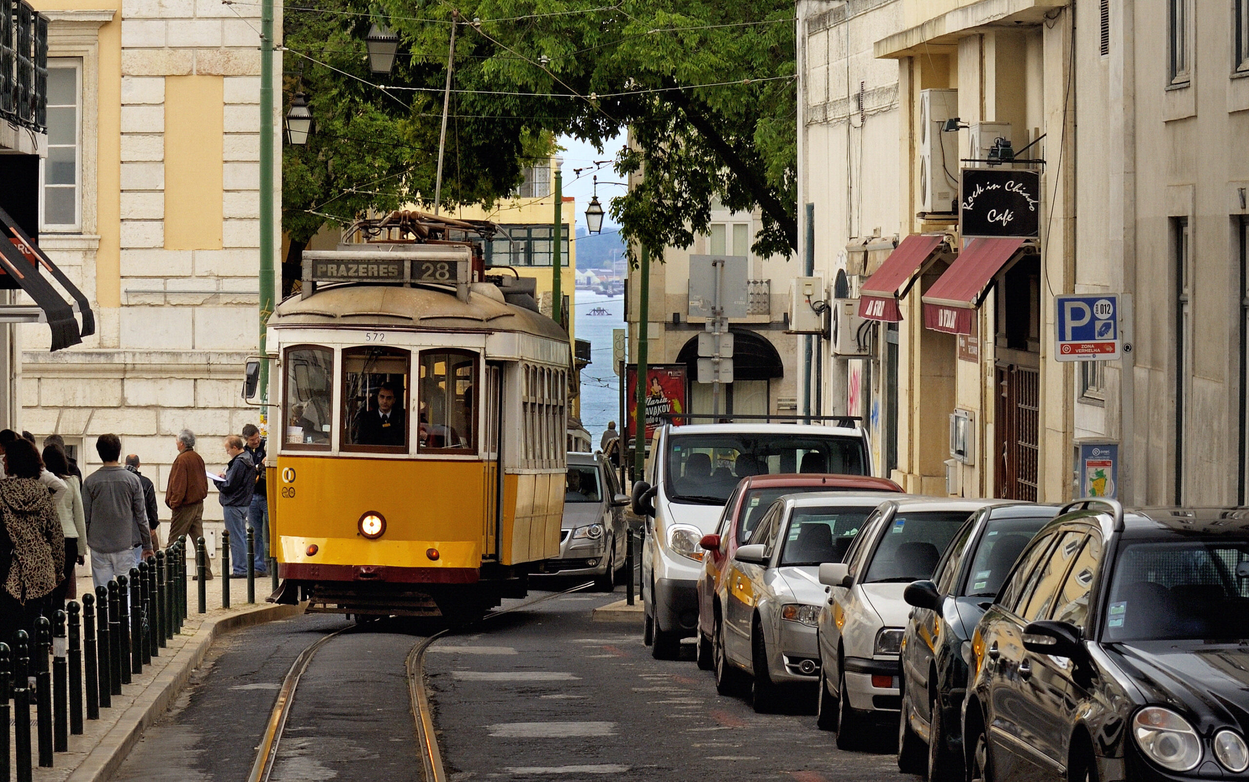 Yellow route 28 tram on a Lisbon street beside pedestrians and a row of parked cars.
